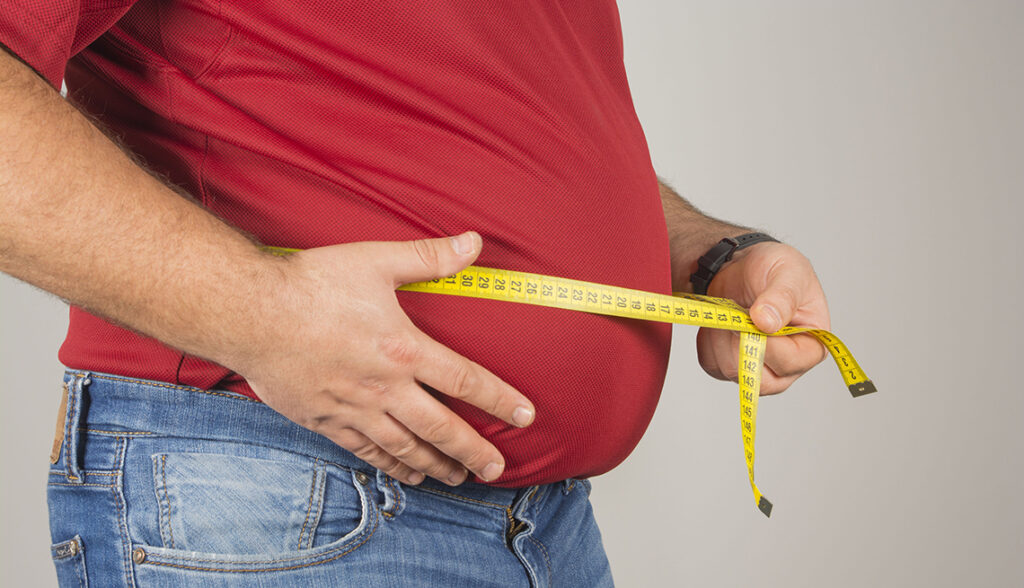 A man with a large belly wearing a red shirt and jeans measuring his waist with a yellow tape measure.