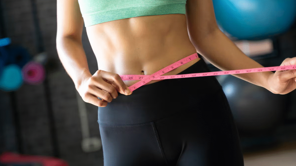A woman in a green sports bra and black leggings measuring her waist with a pink measuring tape in a gym setting.